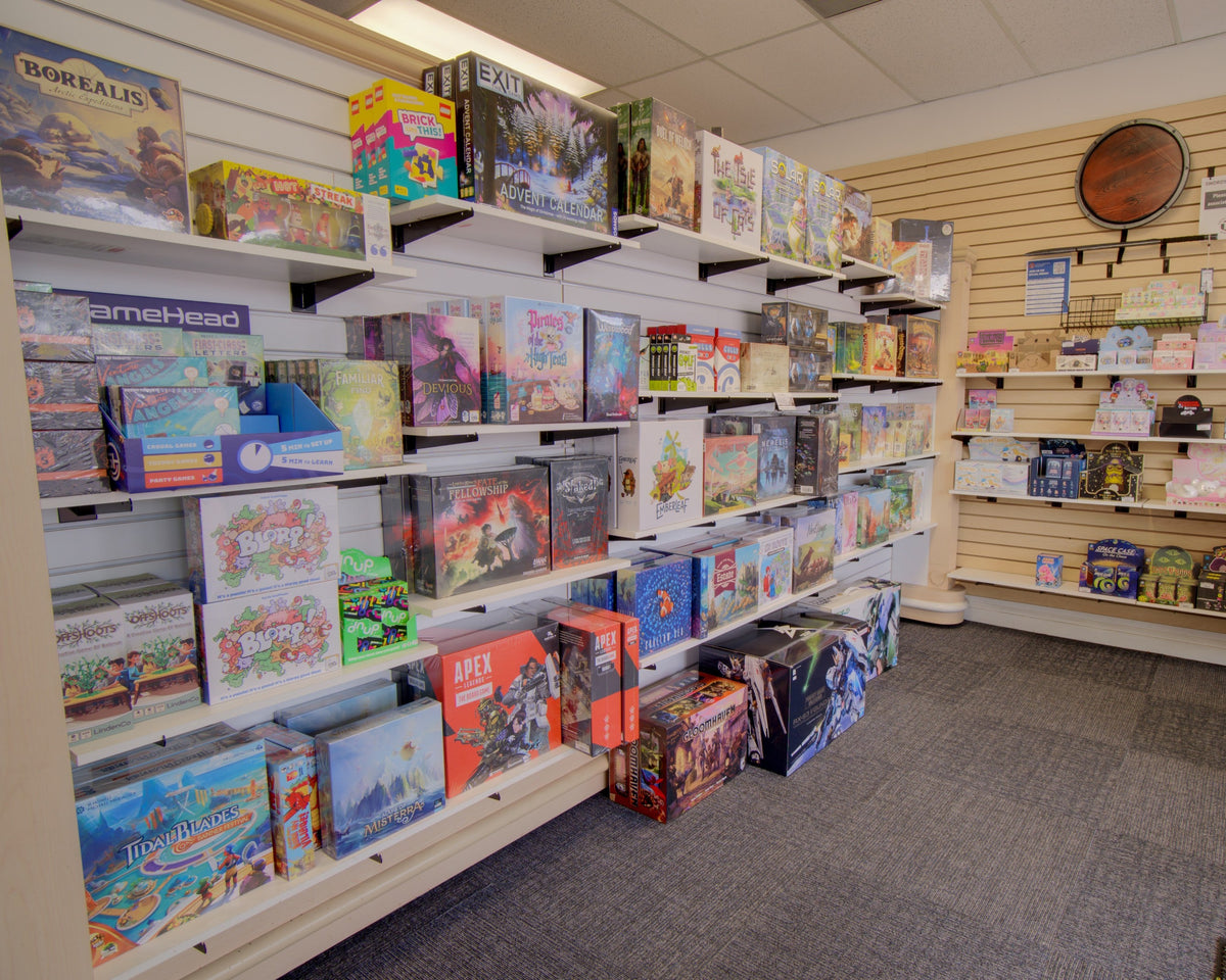 A retail store interior showing shelves filled with board games and boxed toys. The space has light wood slatwall panels, carpeted floor, and fixtures including a round wall clock and hanging swords.